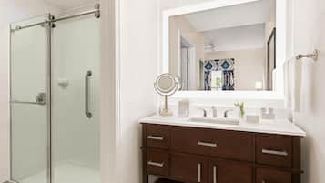 Un baño con cabina de ducha de vidrio, un lavabo blanco con espejo y un mueble de tocador de madera con cajones.