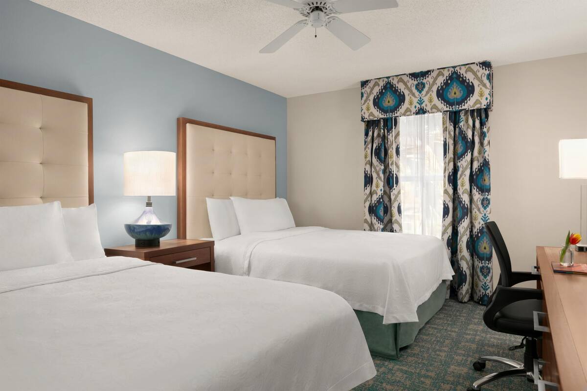 A hotel room with two beds, a ceiling fan, a desk with a chair, and a window with patterned curtains.