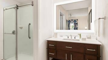 Un baño con cabina de ducha de vidrio, lavamanos blanco y tocador de madera con cajones.