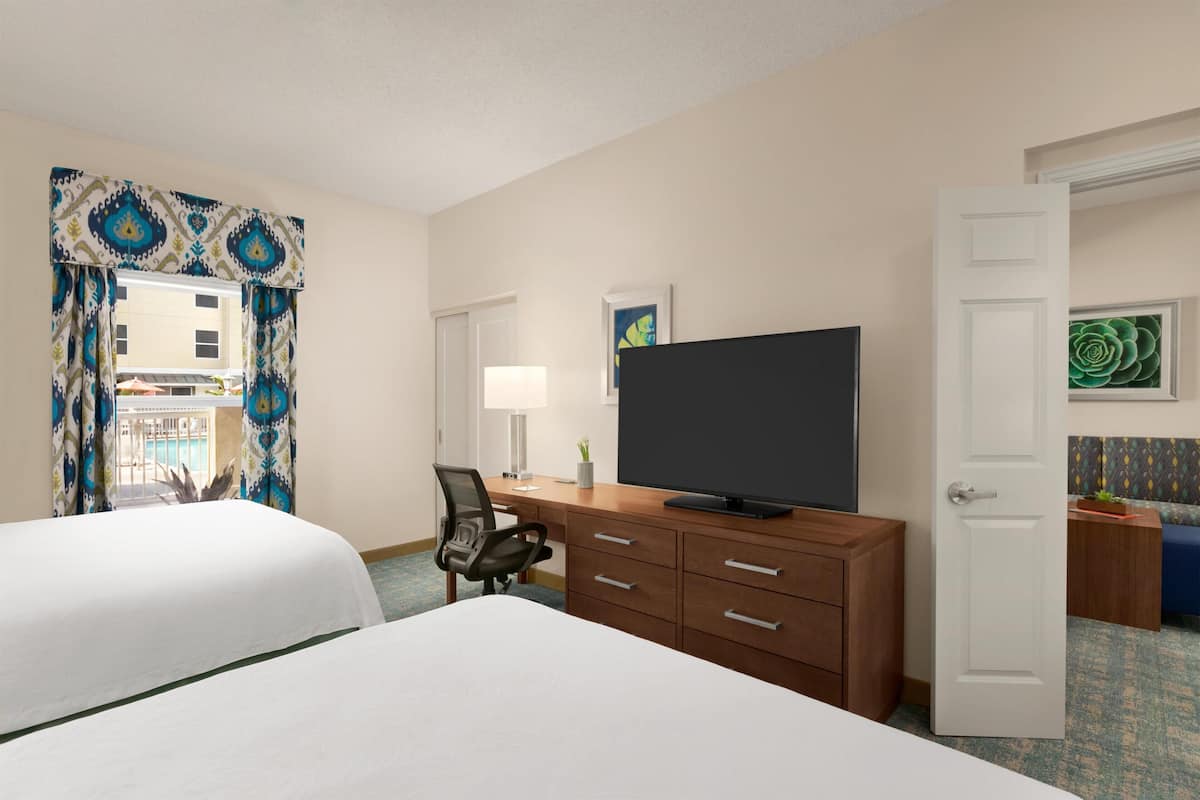 A hotel room with two beds, a desk with a television, a chair, and a door leading to another room.