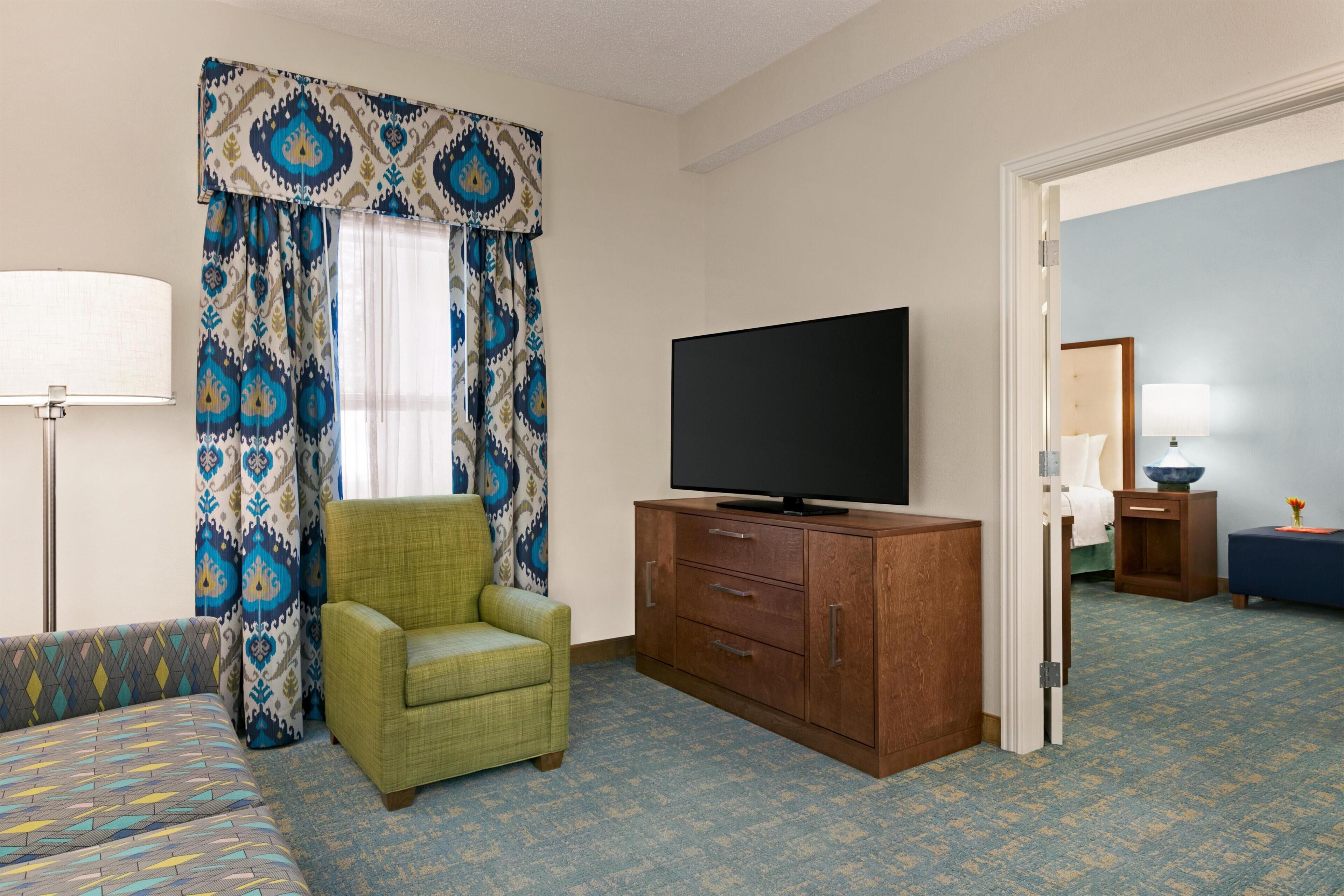 Quarto de hotel com cama, televisão sobre um armário de madeira, poltrona verde, cortinas estampadas e tapete azul.