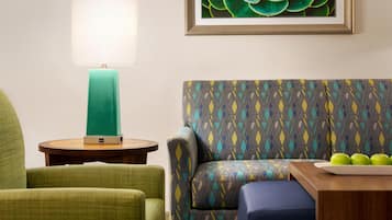 A room with a green armchair, a patterned sofa, a small round table with a teal lamp, and a framed succulent plant.