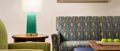 A room with a green armchair, a patterned sofa, a small round table with a teal lamp, and a framed succulent plant.