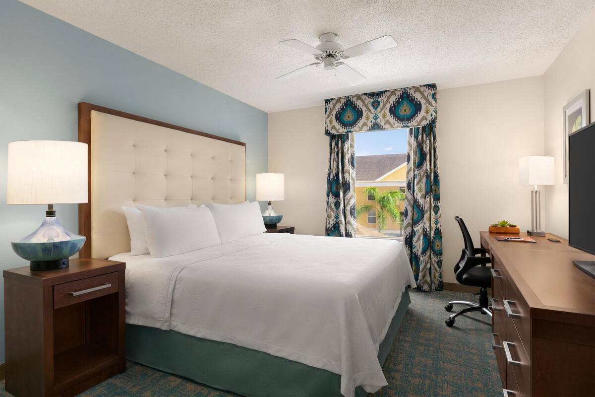 A hotel room with a bed, two bedside tables with lamps, a desk, and a ceiling fan.