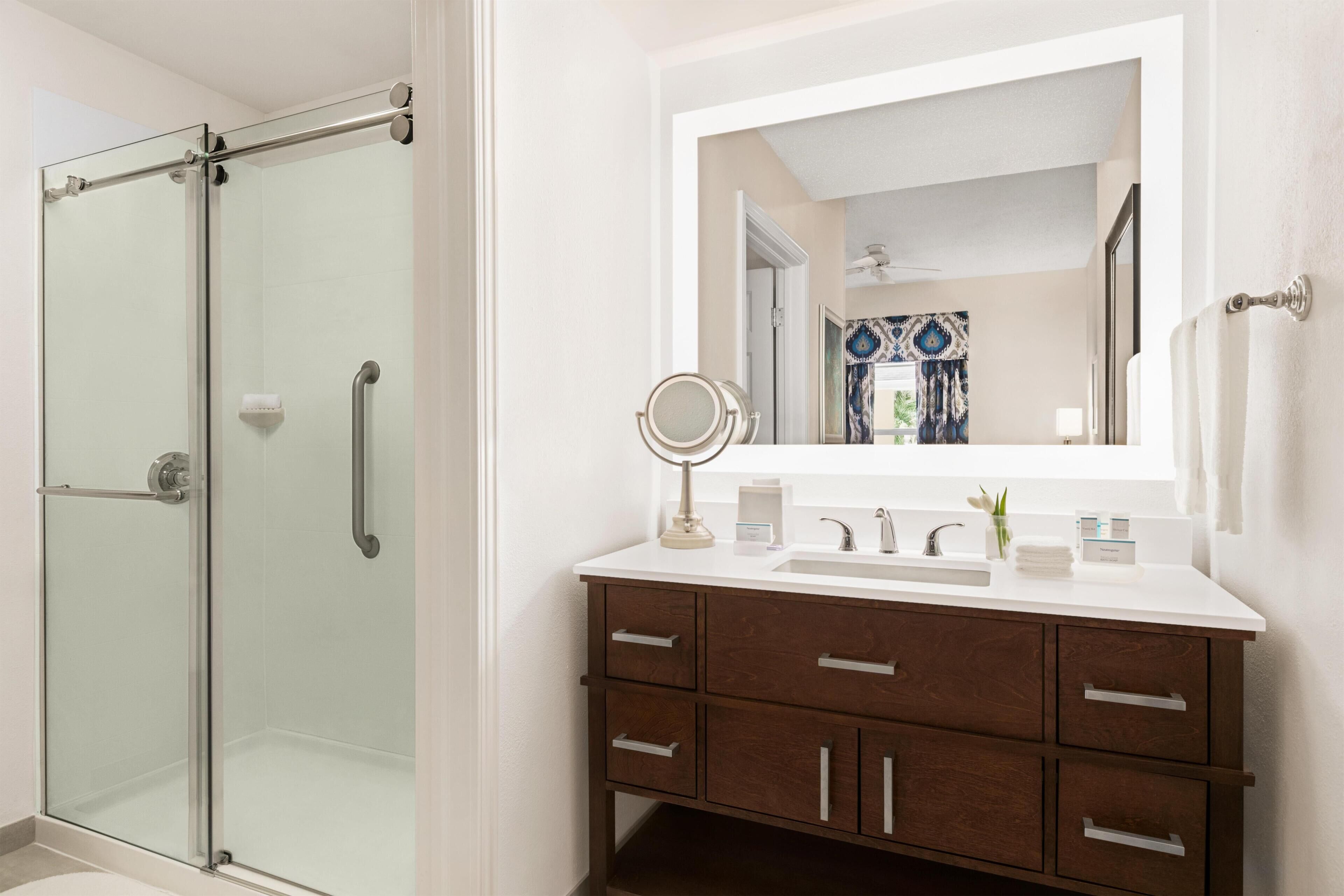 Un baño con cabina de ducha de vidrio, un lavabo blanco con espejo y un mueble de tocador de madera con cajones.