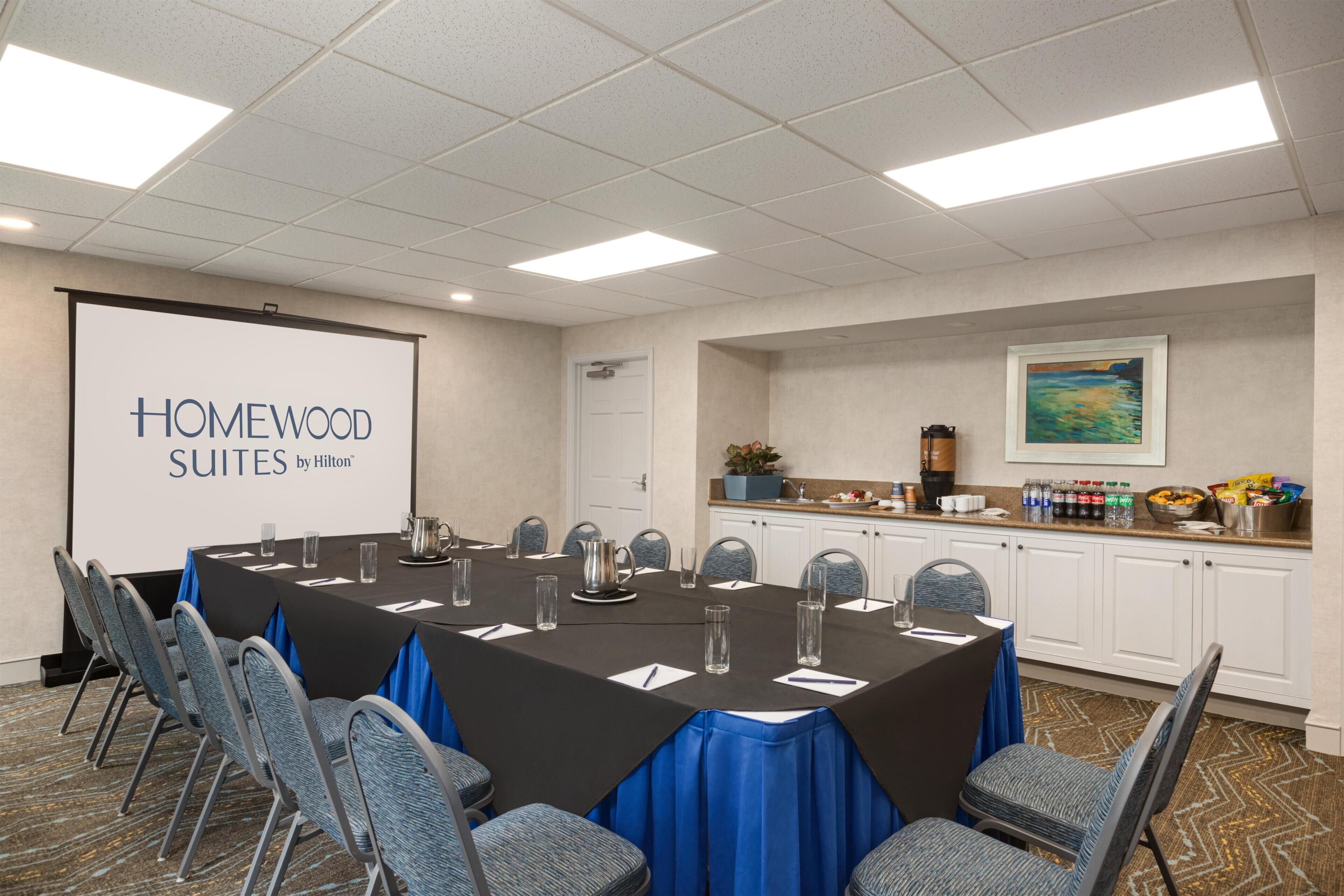 A conference room with a long table, chairs, a projector screen, and a kitchenette area.