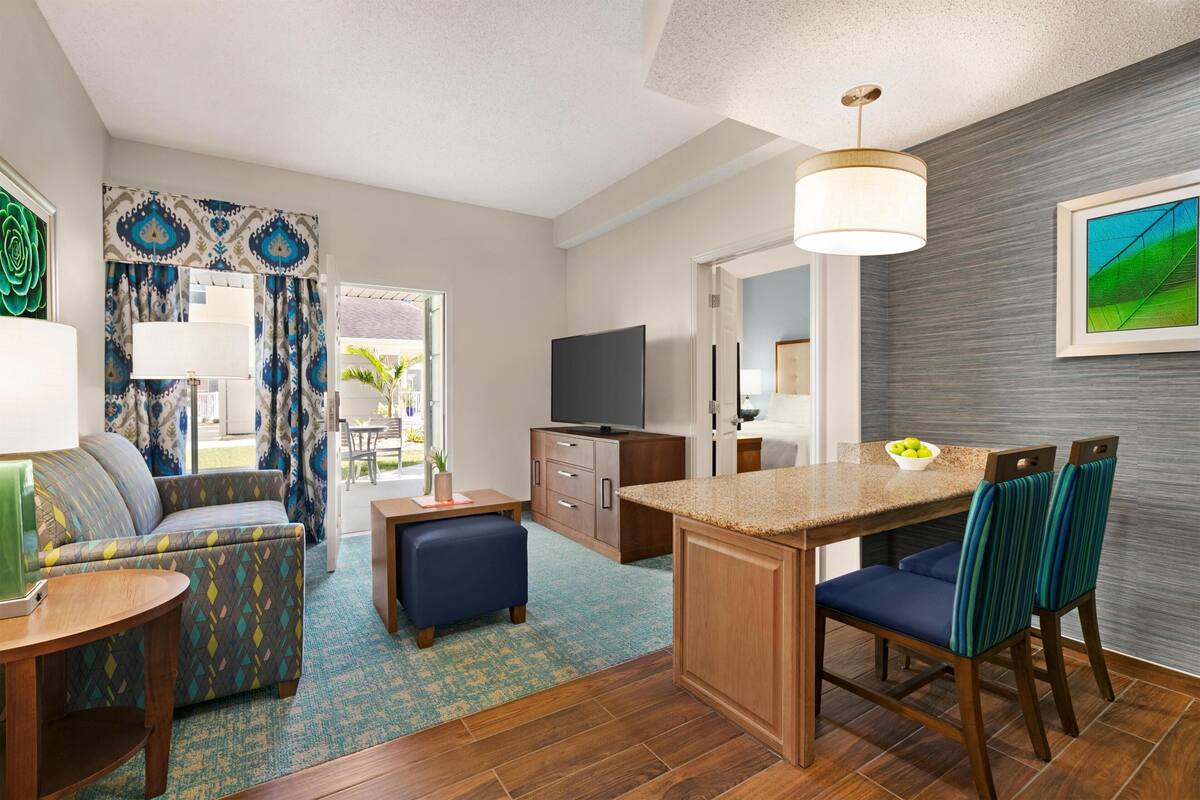 A modern hotel room with a sofa, ottoman, dining table, chairs, a TV, and a kitchen area.