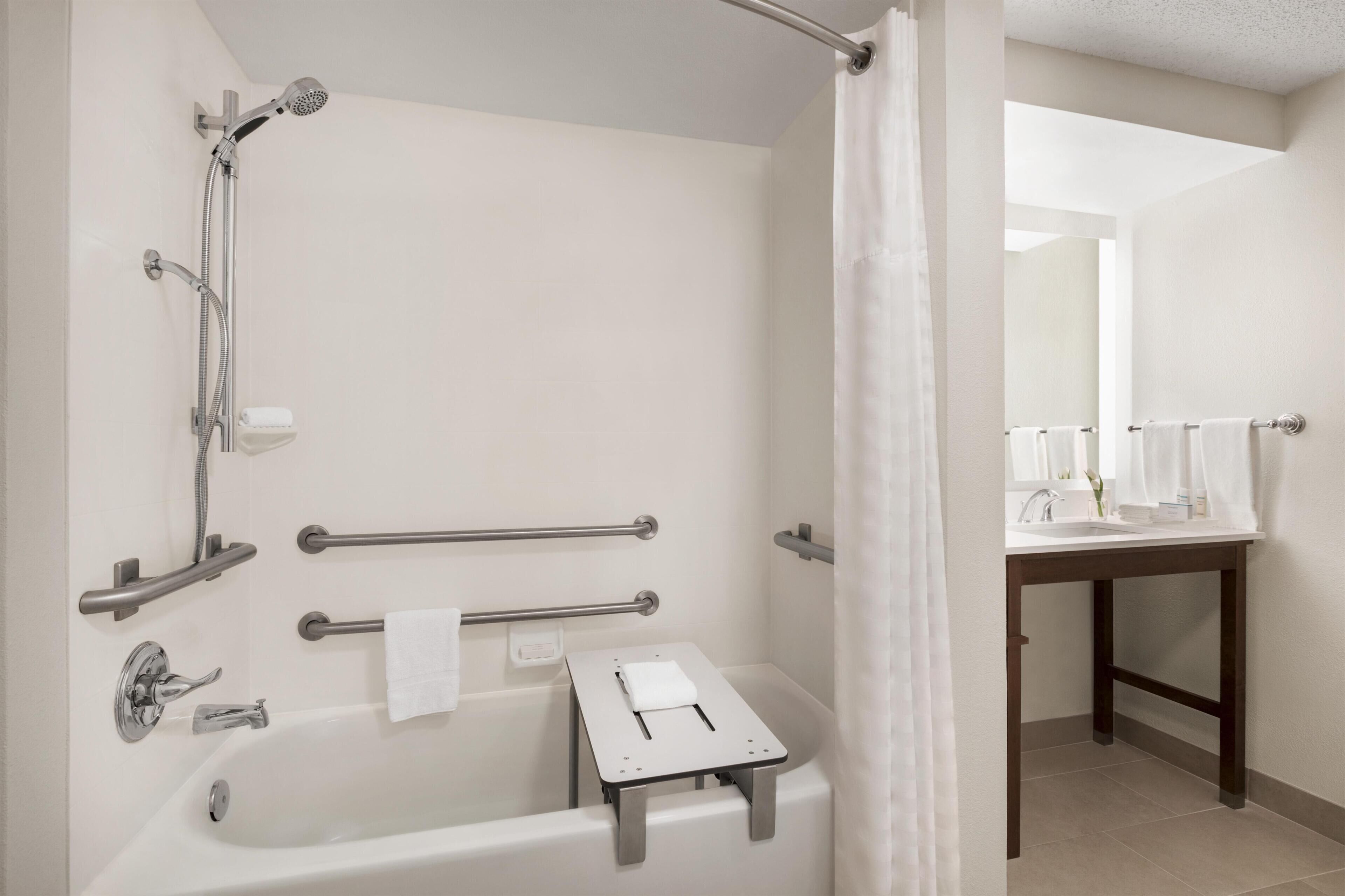 A bathroom with a bathtub, shower, handrails, a small table with a towel, and a sink with a mirror.