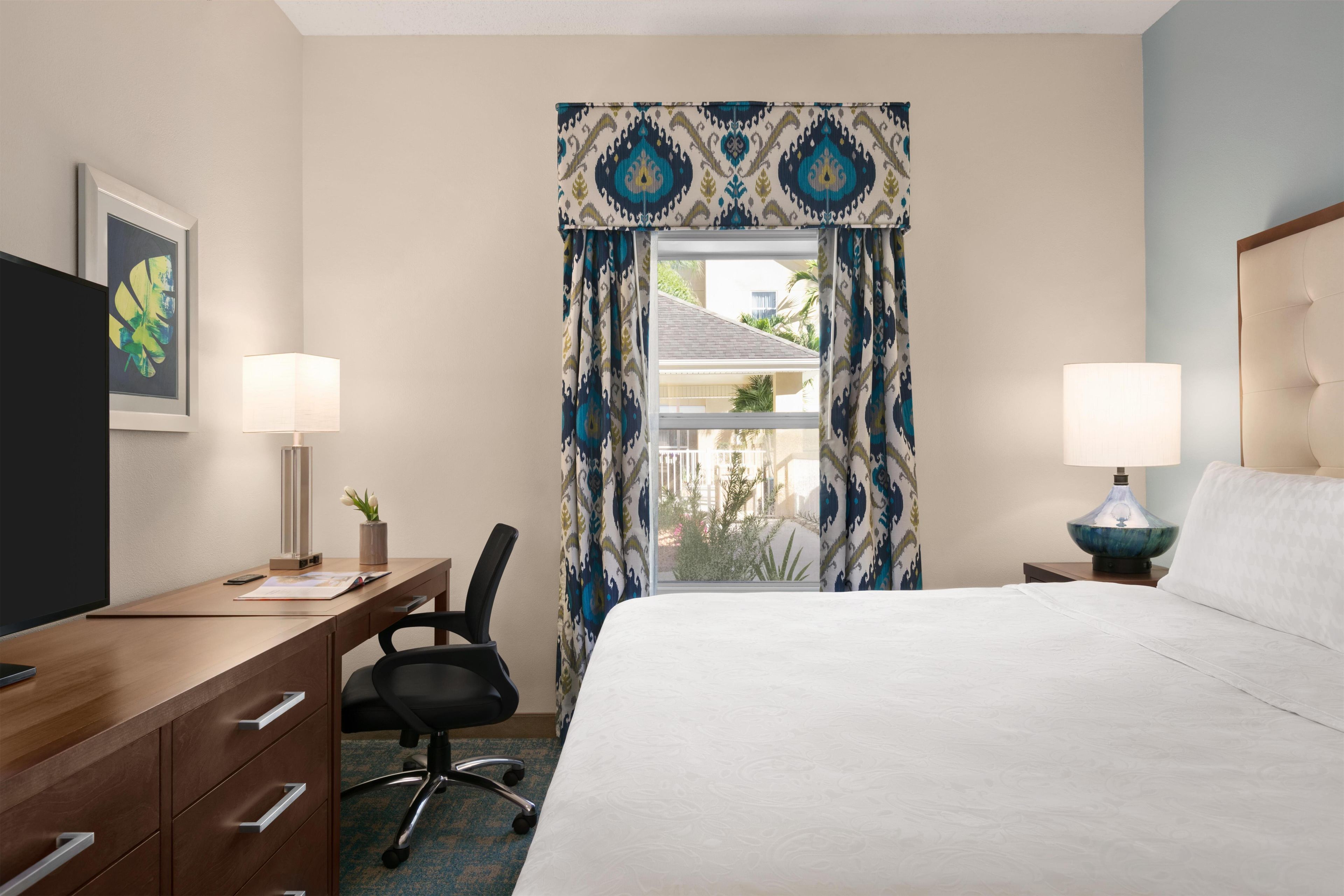 A hotel room with a bed, desk, chair, two lamps, a TV, and a window with curtains.