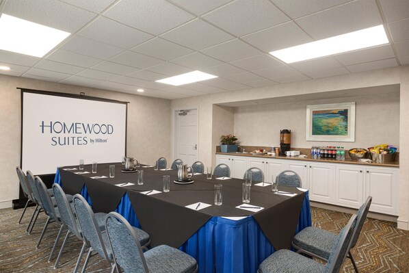 A conference room with a long table, chairs, a projector screen, and a kitchenette area. - Homewood Suites by Hilton - Fort Myers (Fort Myers)