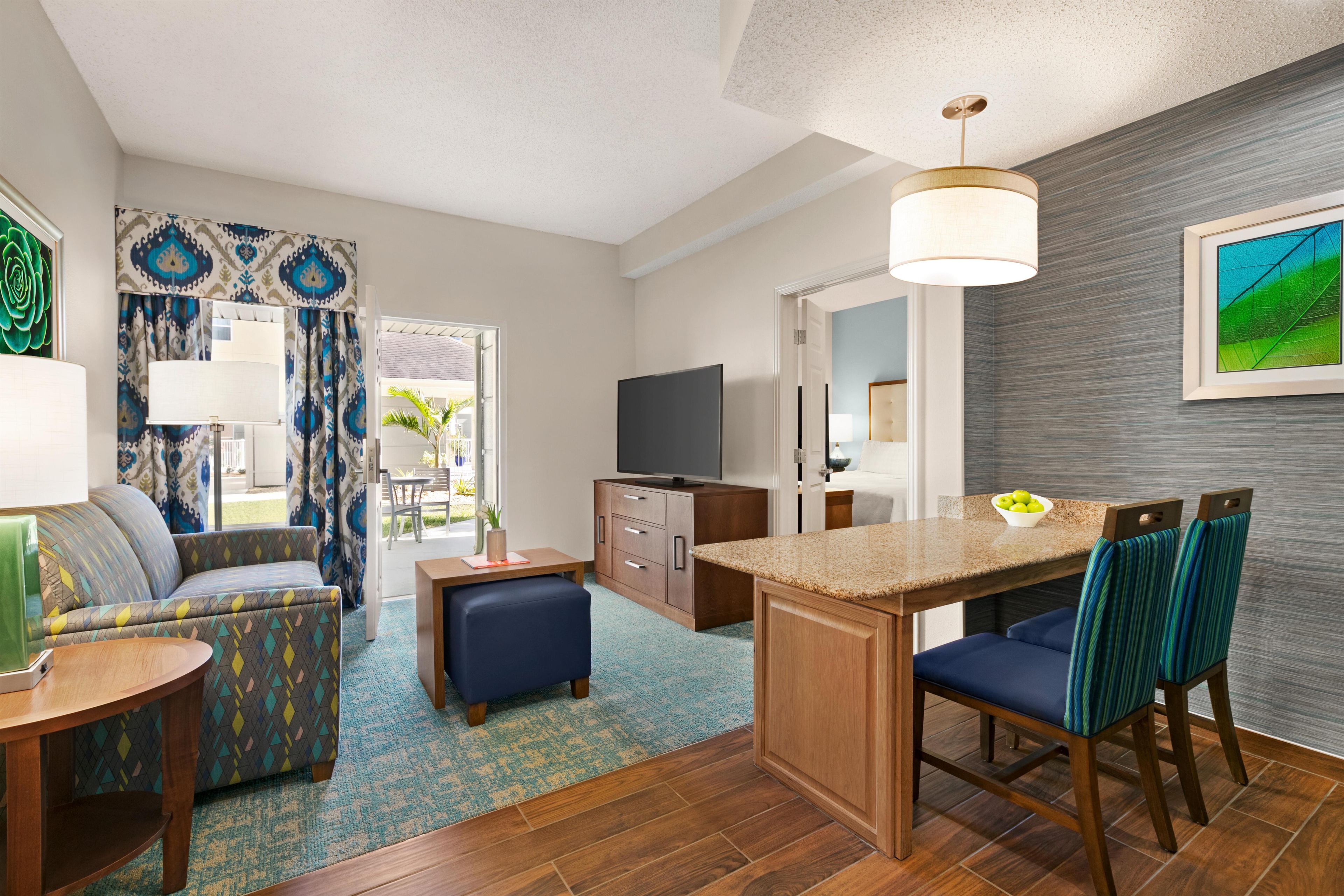 A modern hotel room with a sofa, ottoman, dining table, chairs, a TV, and a kitchen area.