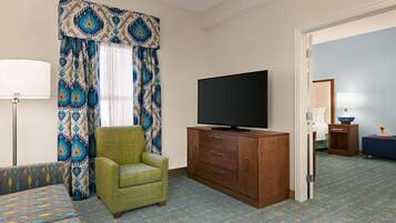 Habitación de hotel con una cama, un televisor sobre un mueble de madera, un sillón verde, cortinas estampadas y una alfombra azul.