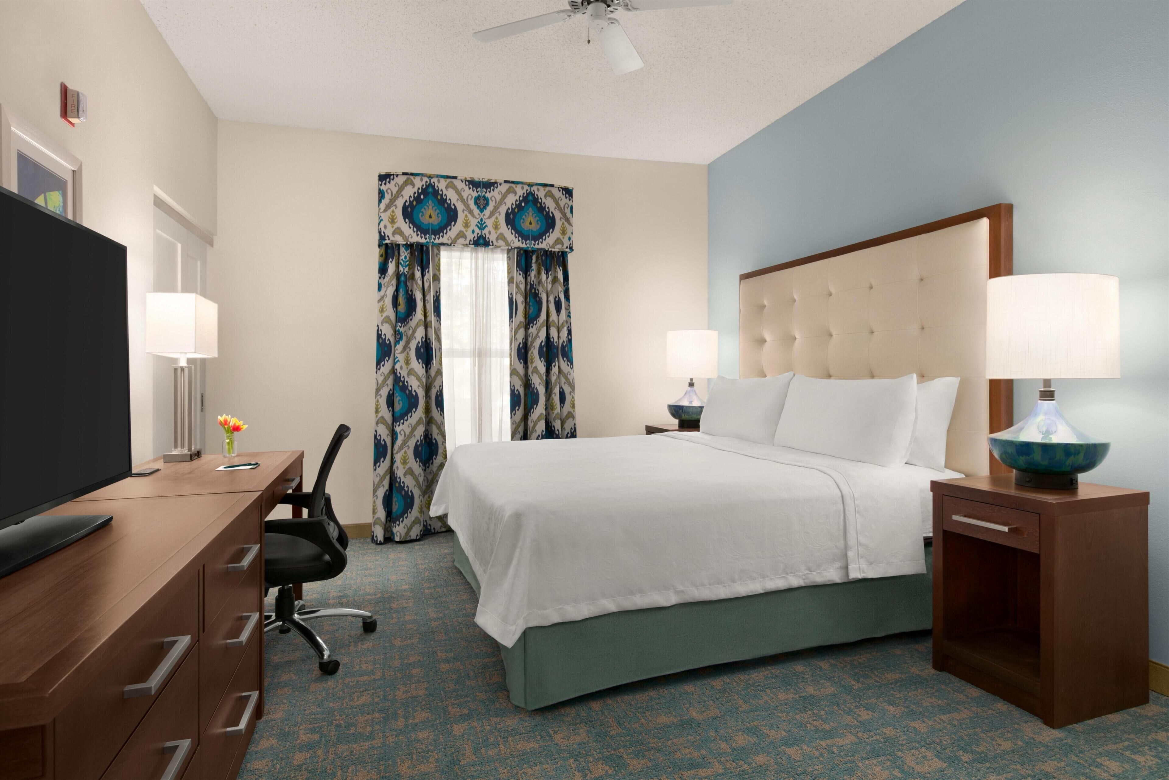 A hotel room with a bed, bedside lamps, a desk with a TV, a chair, and a window with curtains.