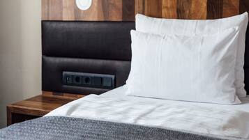 A neatly made bed with white bedding and a grey throw blanket, a wooden headboard with a circular design, and a small bedside table with a power outlet.