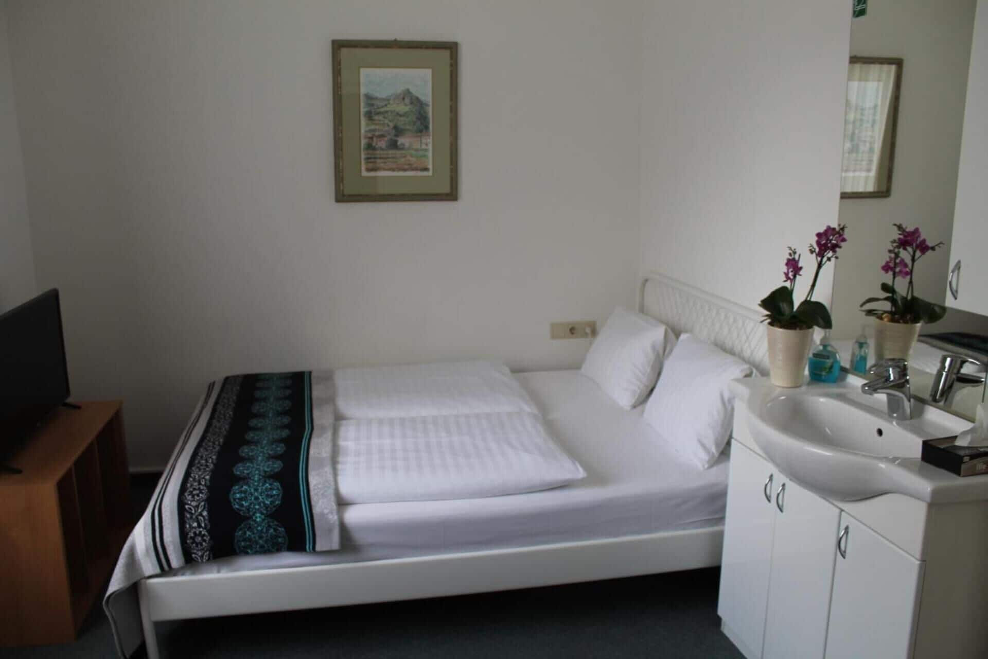 A hotel room with a bed, a bedside table, a TV, a sink, and a mirror.
