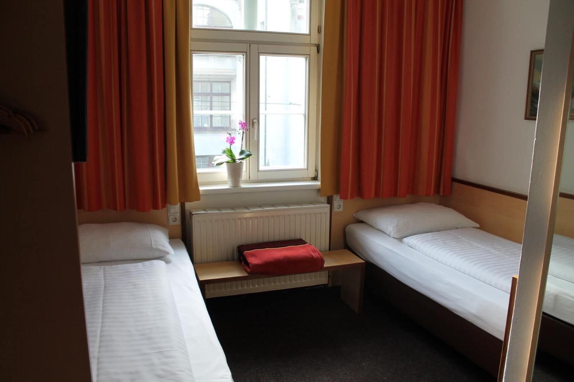 A small, compact room with two single beds, a window with curtains, a radiator, and a small bench with a red cushion.