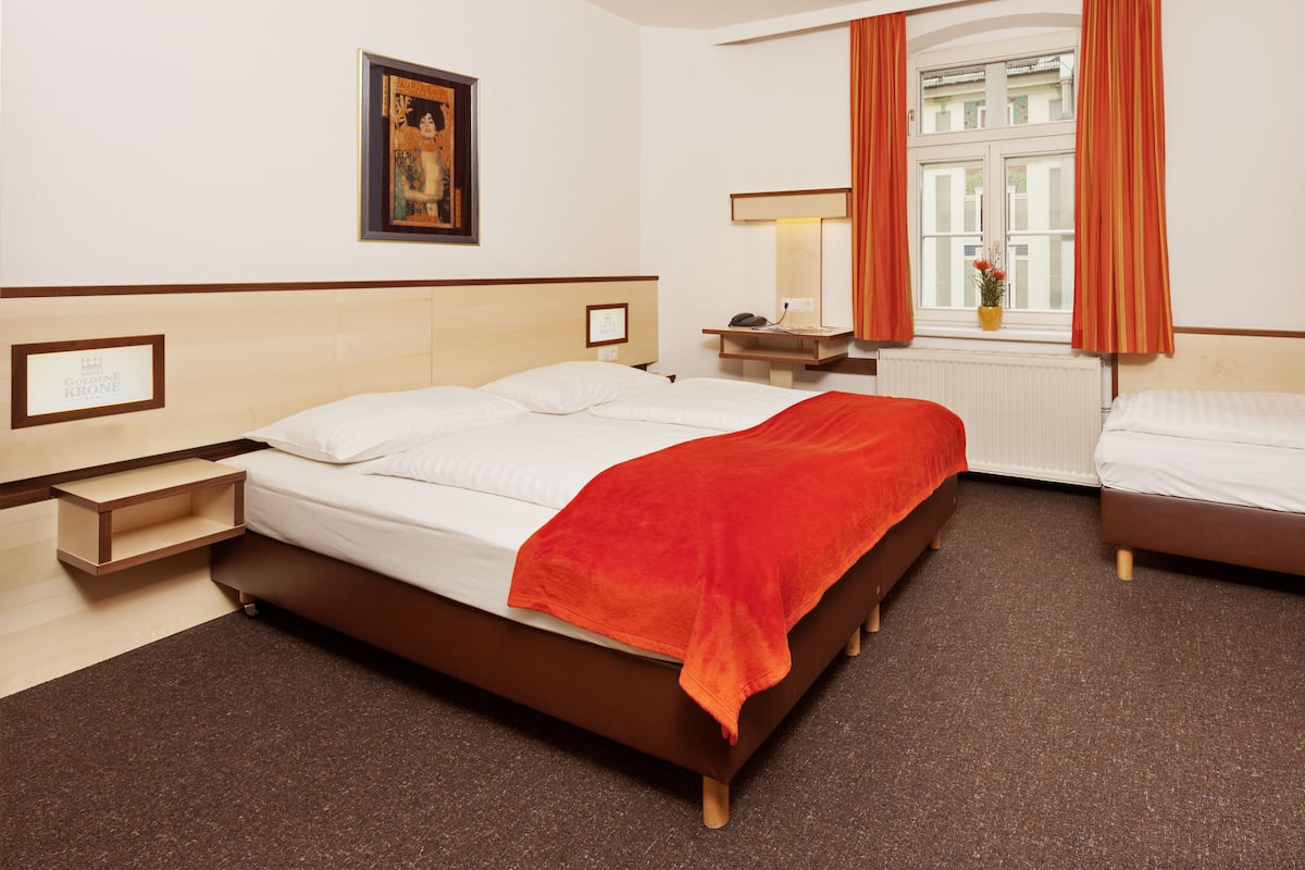 A hotel room with a bed, a bedside table, a nightstand, a window with curtains, and a framed picture on the wall.