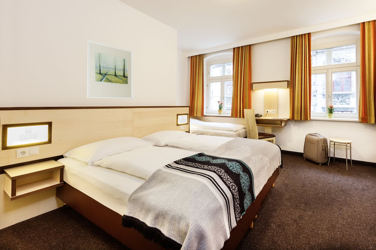 A hotel room with a double bed, a desk, a chair, a small table, a suitcase, and a window with curtains.