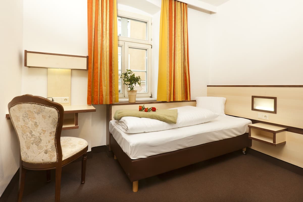 A hotel room with a bed, a desk, a chair, a bedside table, a lamp, and a window with curtains.