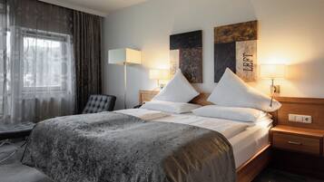Una cama bien hecha con colcha gris texturizada, dos lámparas de noche, un cabecero de madera y una pared con dos cuadros enmarcados.