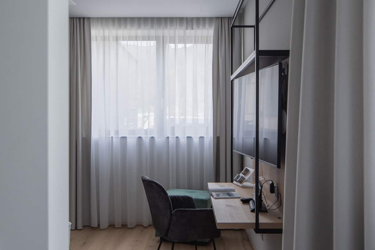 A modern room with a wooden floor, a desk with a chair, a flat-screen TV mounted on the wall, and a window with sheer curtains.