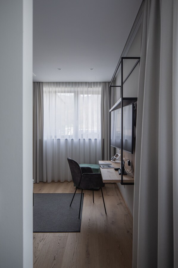 A modern room with a wooden floor, a desk with a chair, a flat-screen TV mounted on the wall, and a window with sheer curtains. - Hotel Goldenes Schiff (Bad Ischl)