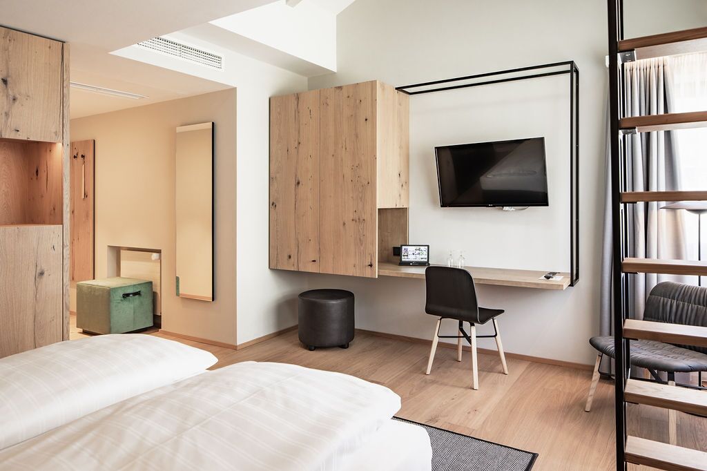 A modern hotel room with a large bed, a desk with a chair, a TV mounted on the wall, and a wooden wardrobe.