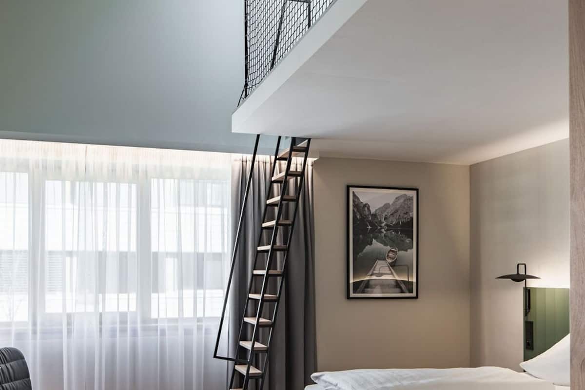 A bedroom with a bed, a chair, a ladder, a framed picture on the wall, and a window with curtains.