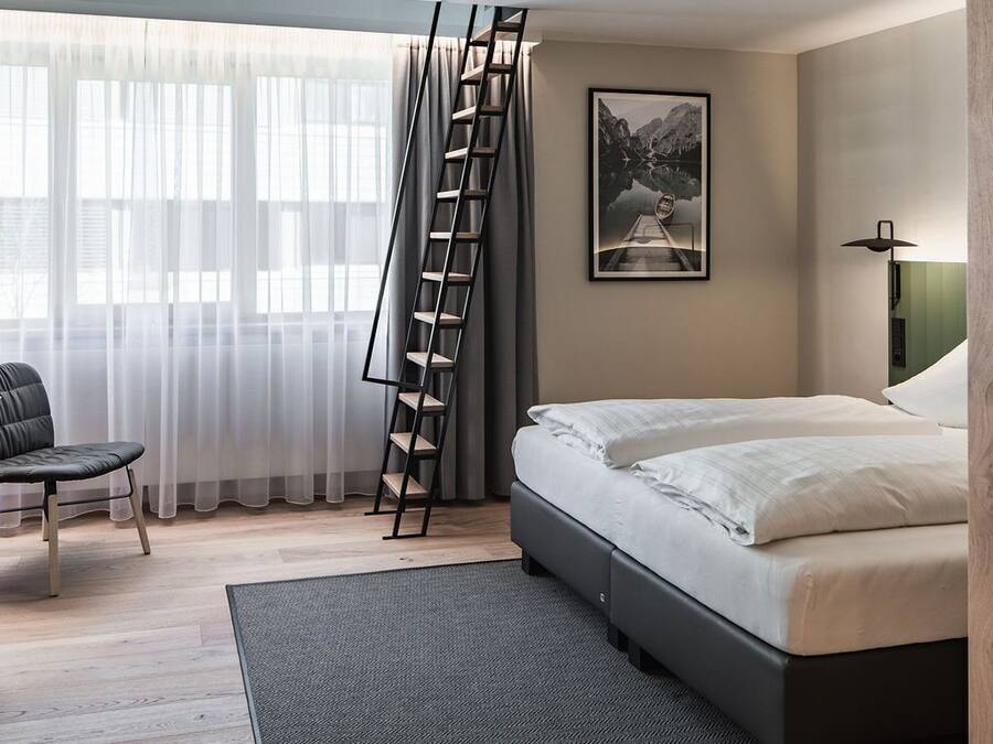 Un dormitori amb un llit, una cadira, una escala, un quadre a la paret i una finestra amb cortines.
