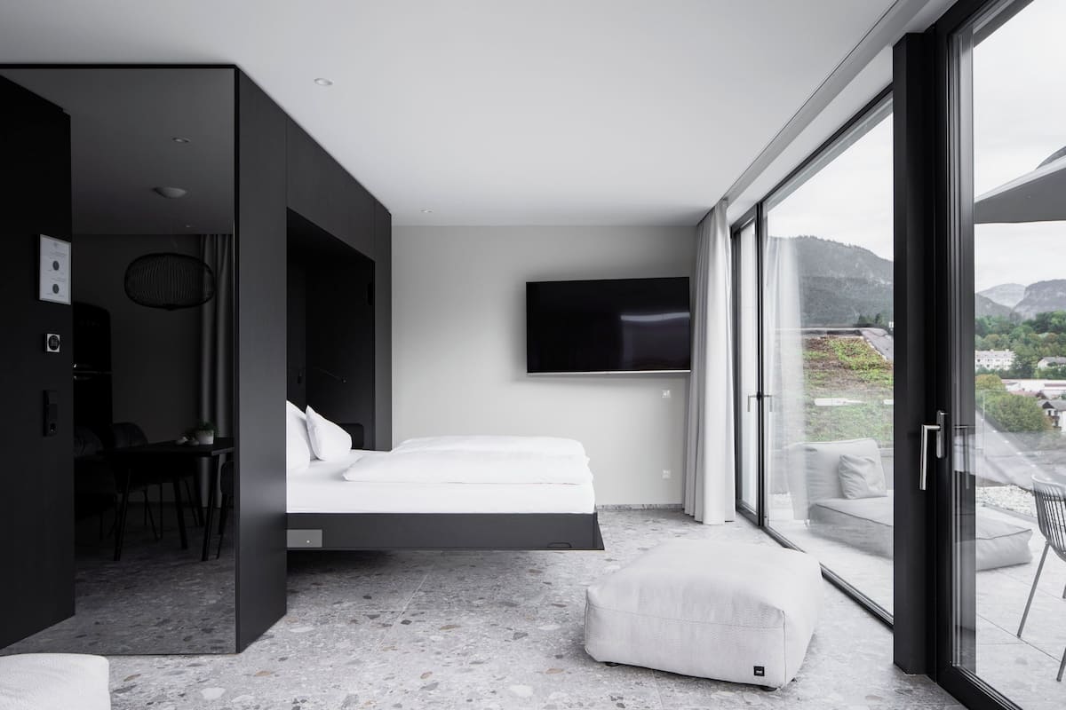 A modern hotel room with a bed, a TV, a desk, and a balcony with a view of the mountains.