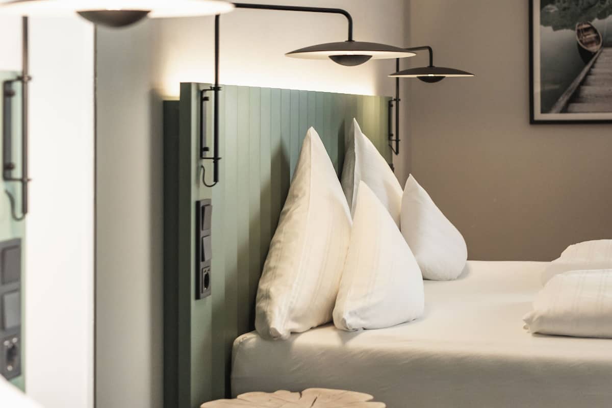 A hotel room with a bed, white pillows, a wooden side table, and a wall-mounted lamp.