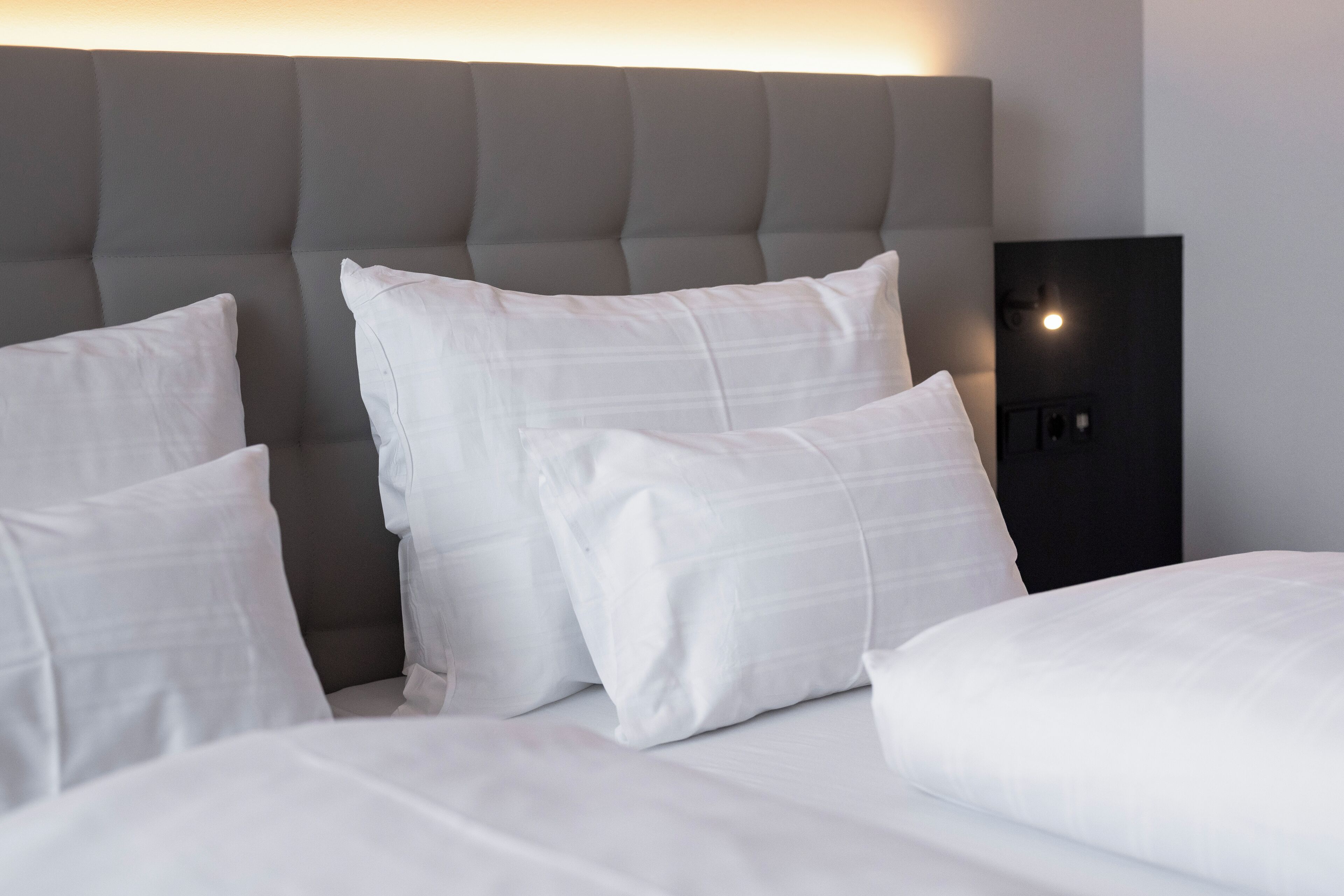 Ein ordentlich bezogenes Bett mit weißer Bettwäsche und Kissen, ein grauer möbelpolsterter Kopfteil und ein Nachttisch in Schwarz mit einer Lampe.