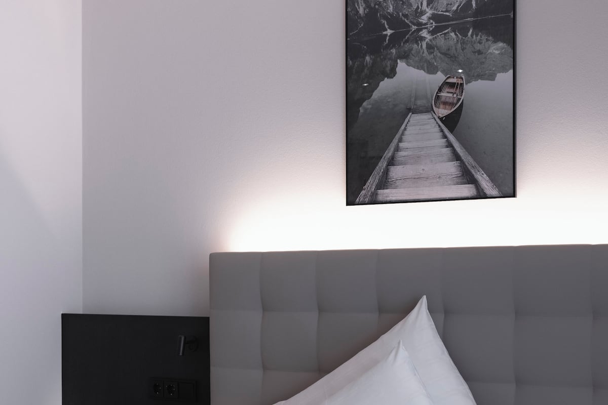A modern hotel room with a large bed, a headboard, a bedside table, and a wall-mounted picture of a mountain landscape.