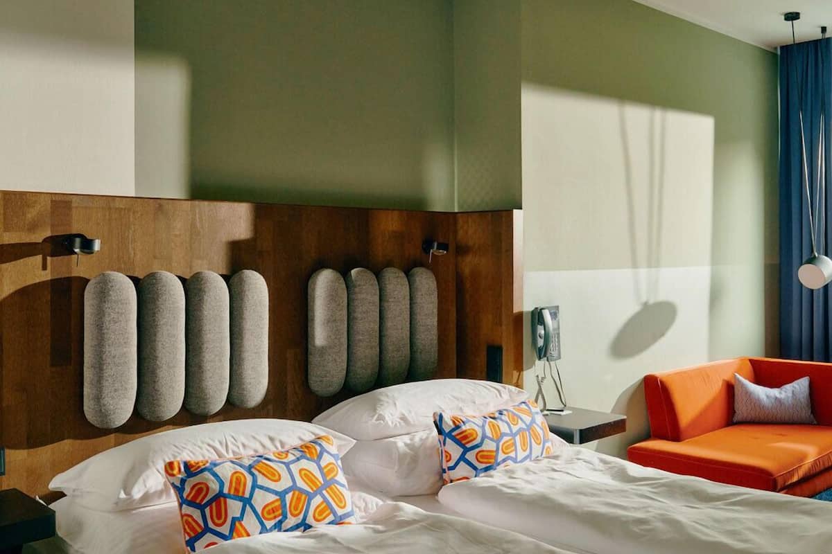 A hotel room with a wooden headboard, a bed with white linens and patterned pillows, an orange armchair, and a bedside table with a lamp.
