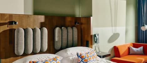 A hotel room with a wooden headboard, a bed with white linens and patterned pillows, an orange armchair, and a bedside table with a lamp.