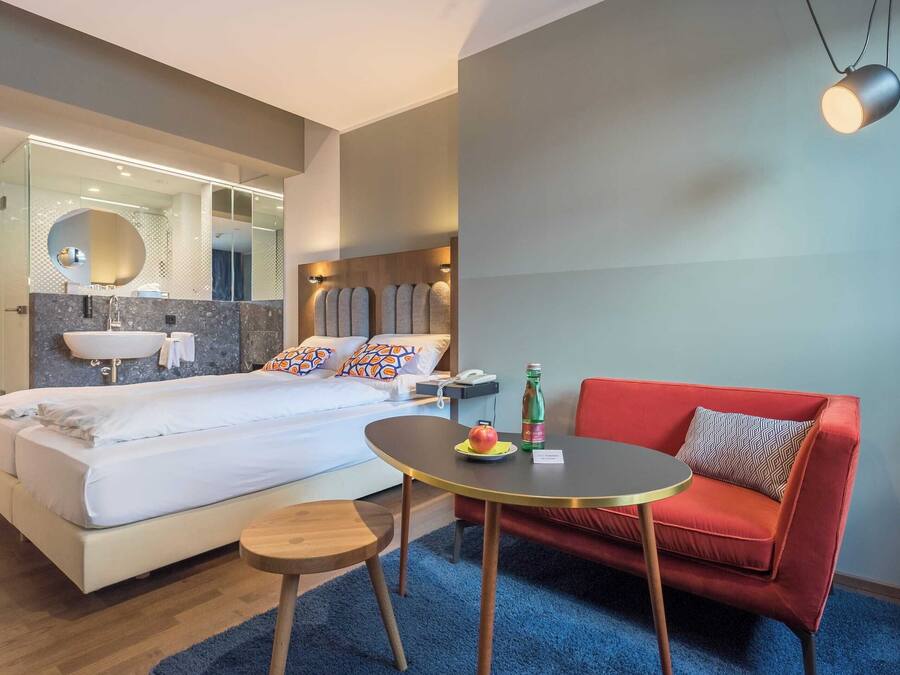 Ein modernes Hotelzimmer mit einem großen Bett, einem roten Sofa, einem kleinen runden Tisch und einem Spiegel über dem Bett.