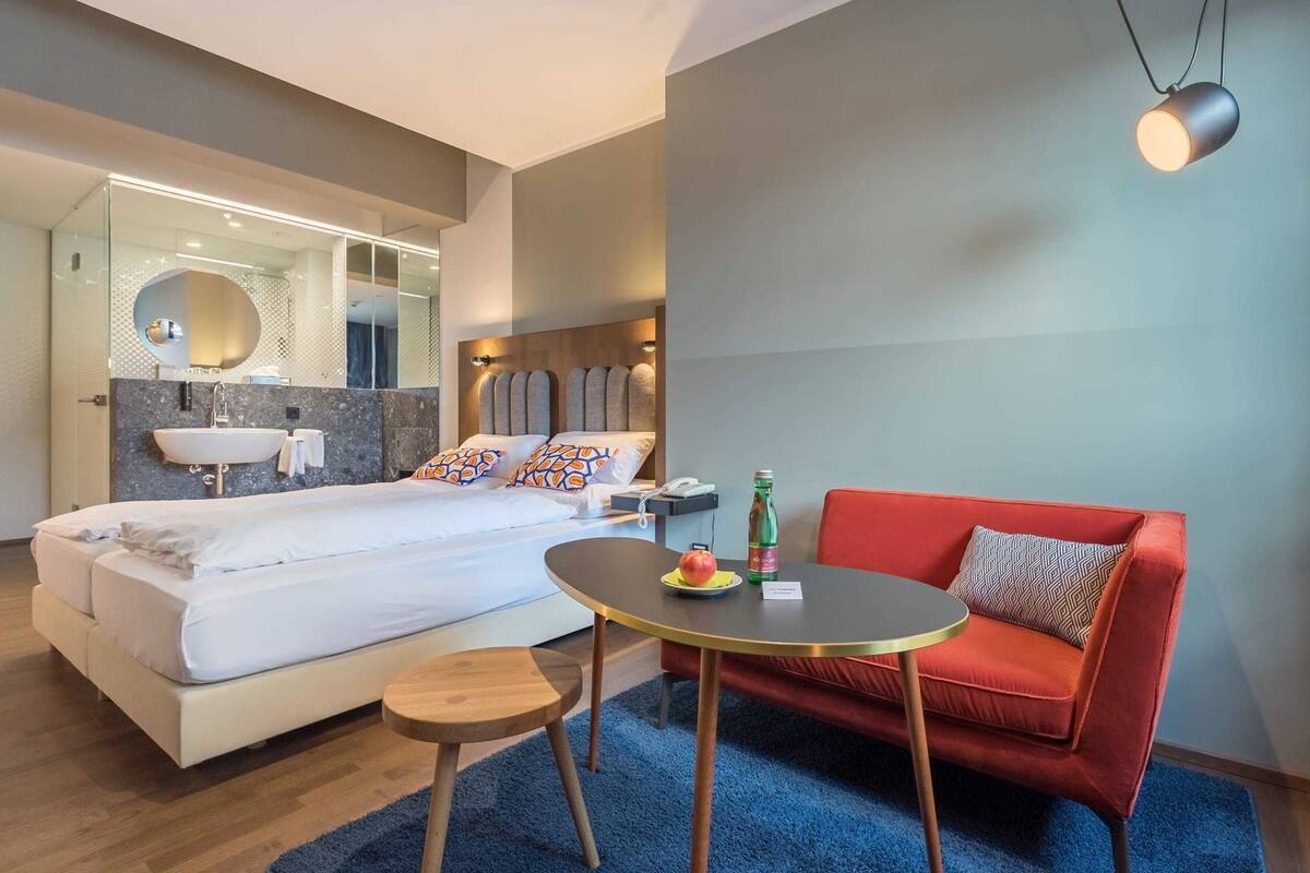 A hotel room with a bed, a round table, a red sofa, a small stool, a mirror, and a wall-mounted light fixture.