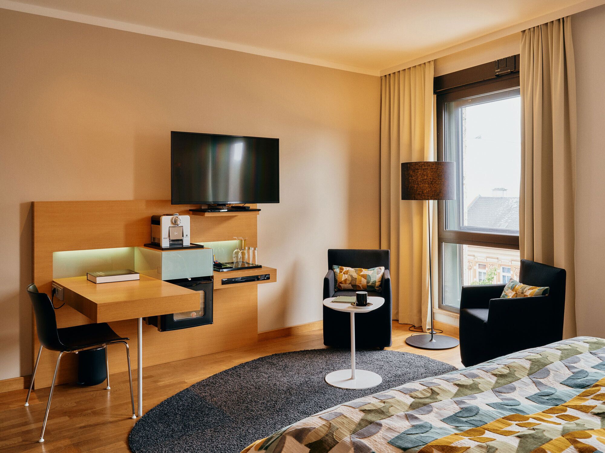 A hotel room with a bed, a desk with a television, a chair, a small table, and a lamp.