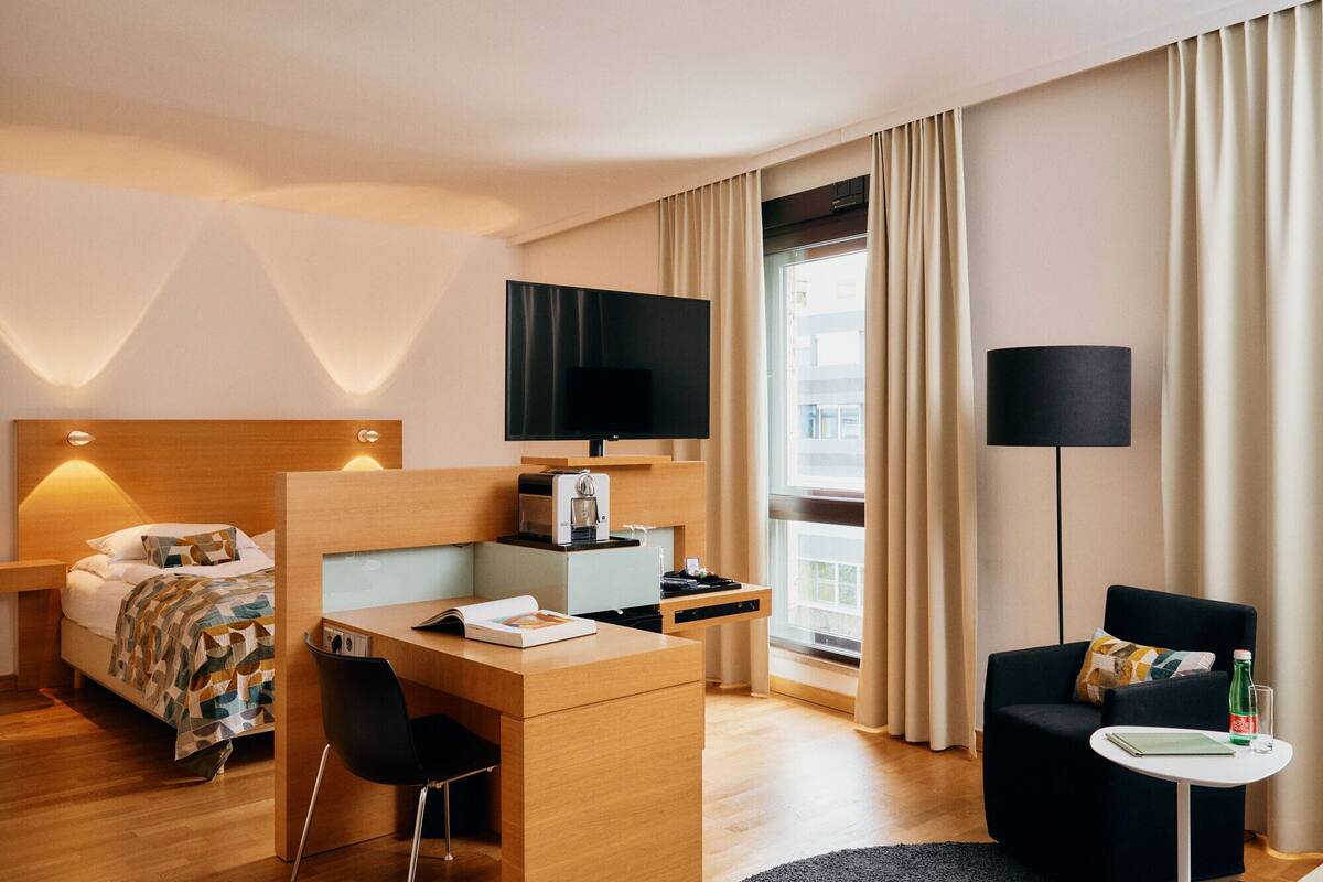 A hotel room with a bed, a desk with a coffee machine, a TV, a sofa, and a window with curtains.