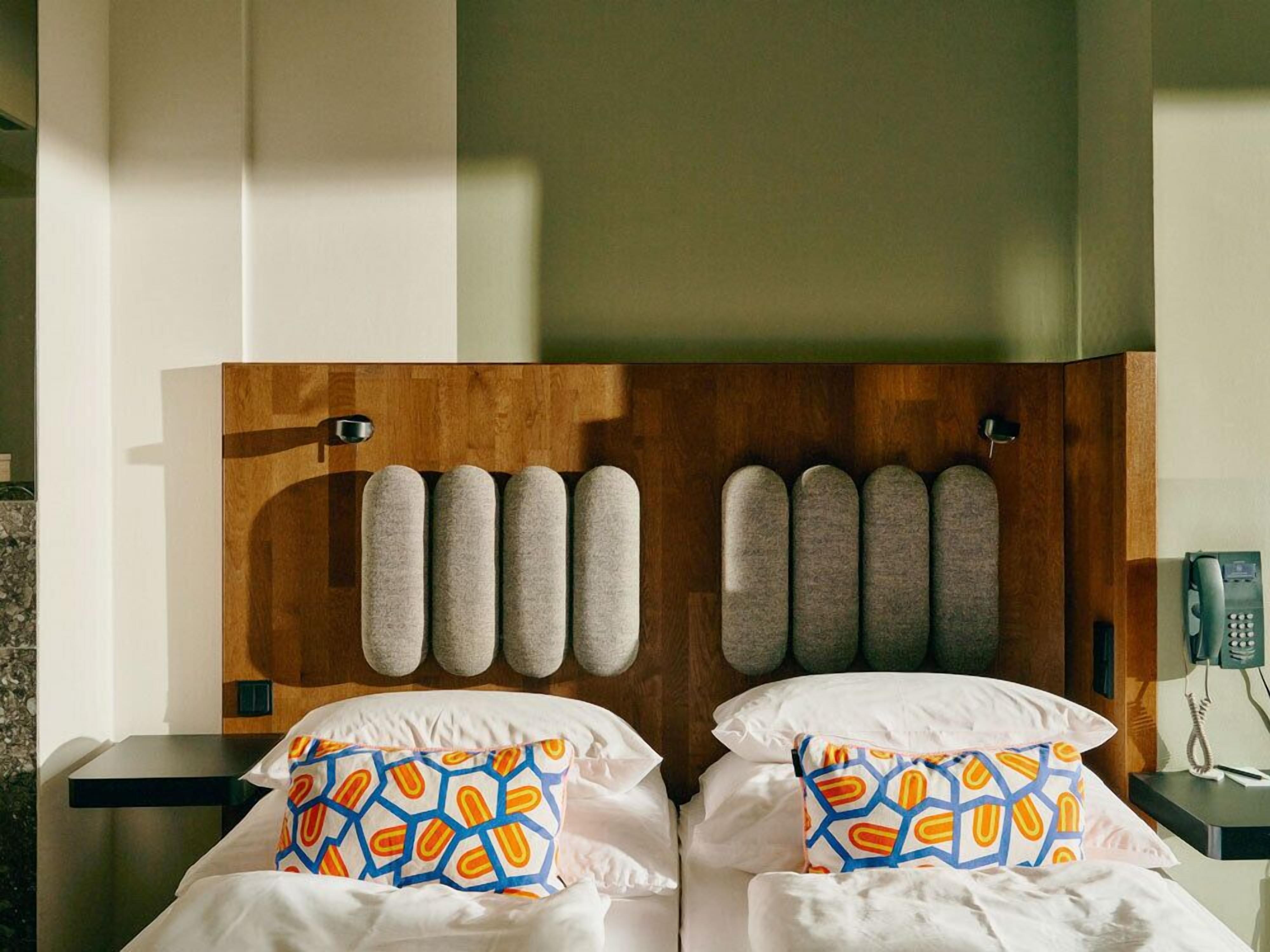 Une chambre d’hôtel avec une tête de lit en bois, deux lits avec du linge de lit blanc et des coussins aux motifs colorés.