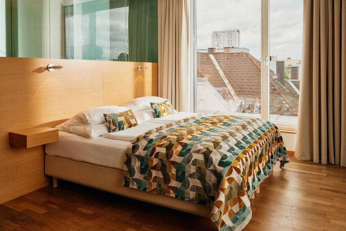 A modern hotel room with a large bed, a wooden headboard, a glass partition, and a view of the cityscape through large windows.