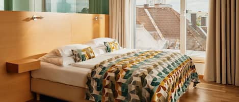 Quarto de hotel moderno com uma cama grande, cabeceira de madeira, divisória de vidro e vista para a paisagem urbana através de amplas janelas.