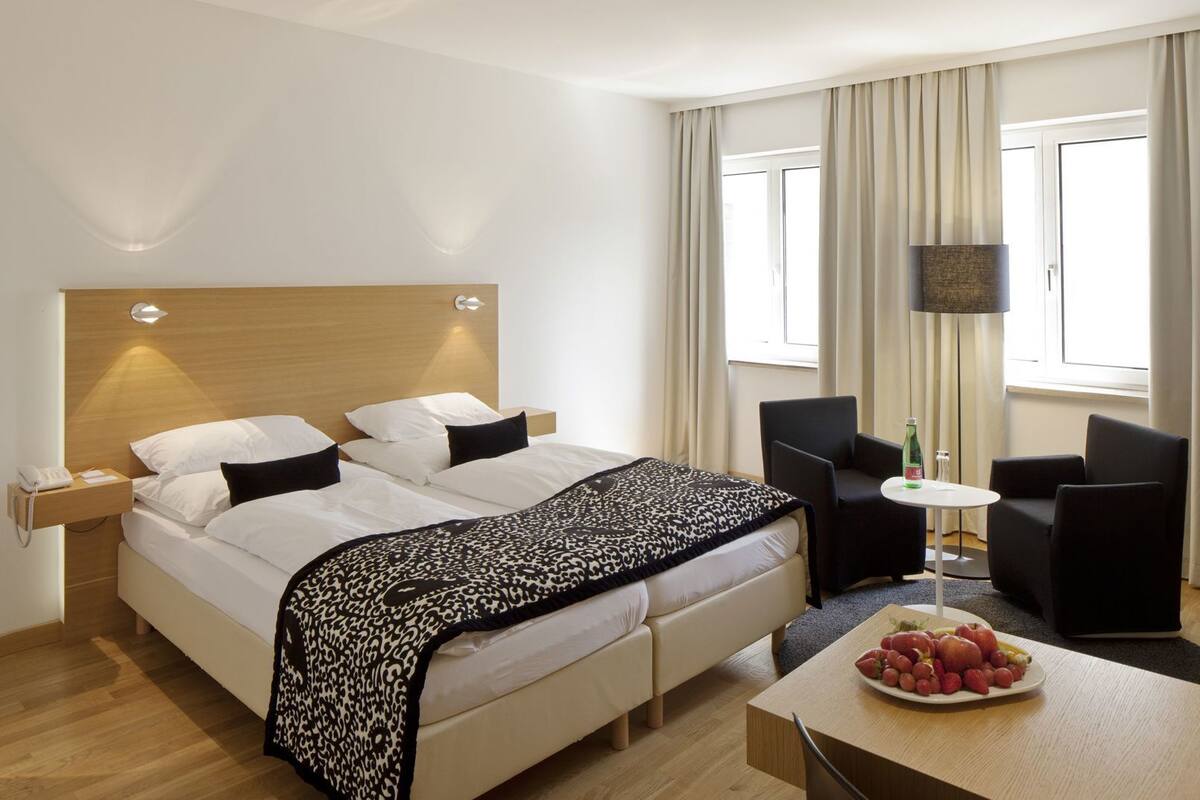 A hotel room with a bed, bedside table, lamp, chair, table, and a fruit plate.