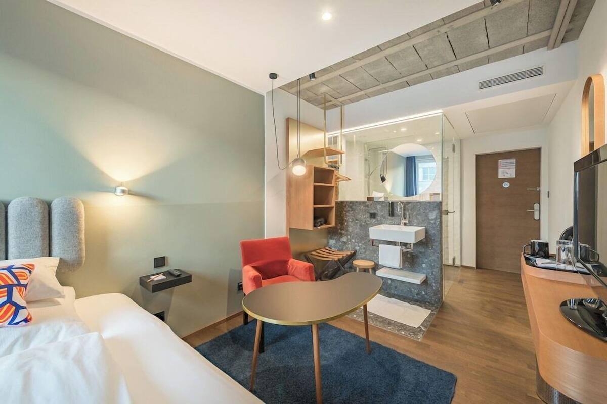 A hotel room with a bed, a desk, a chair, a mirror, a sink, and a television.
