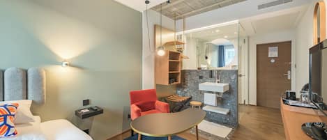 A hotel room with a bed, a desk, a chair, a small table, a mirror, a sink, and a television.