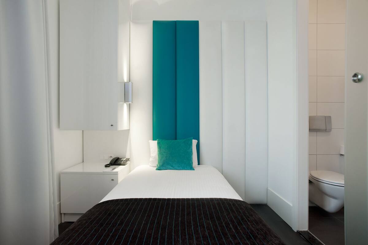 Una habitación de hotel moderna con cama, mesita de noche, baño con inodoro y lavamanos, y un cabecero color aguamarina.