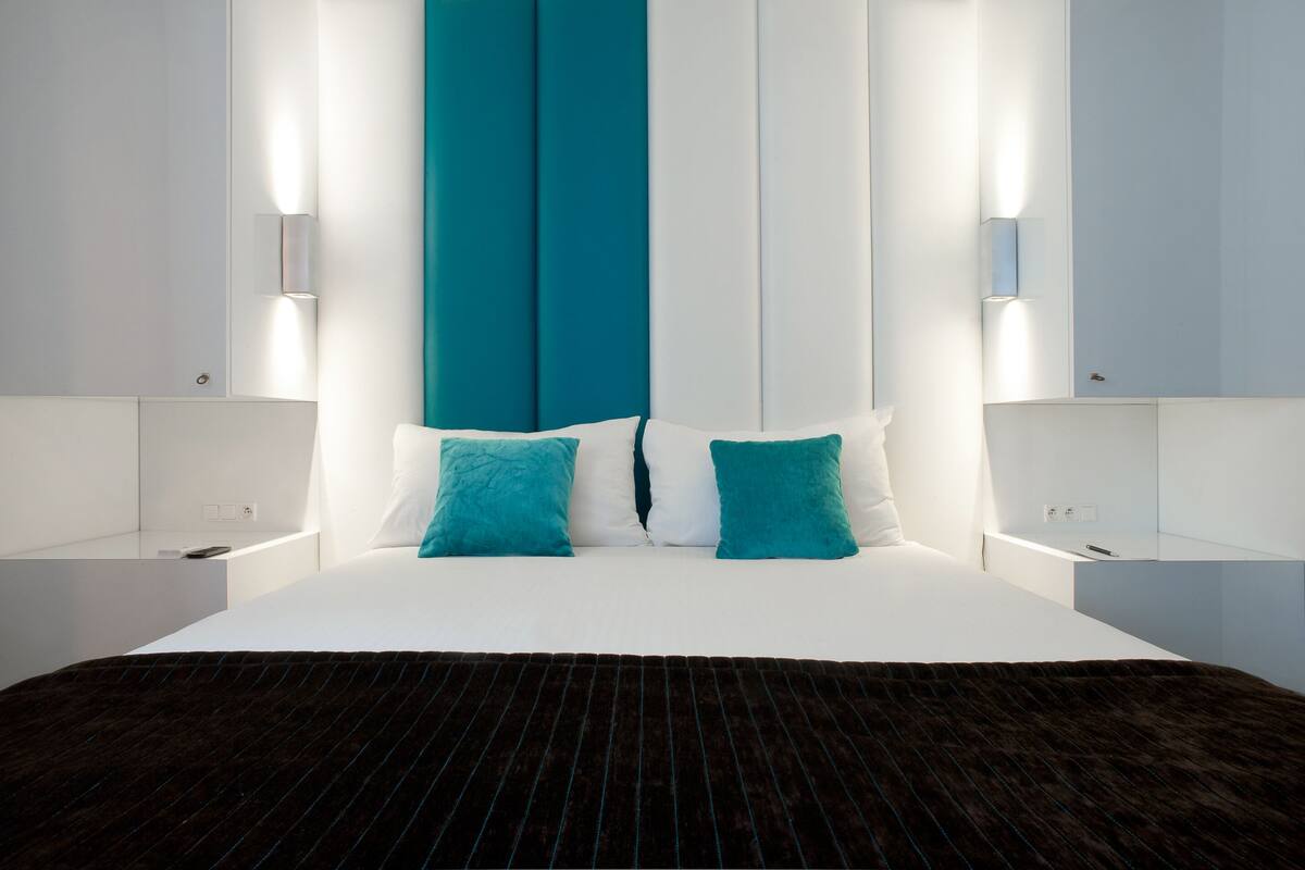 Una habitación de hotel moderna con una cama grande, dos almohadas color turquesa y una manta rayada marrón oscuro.