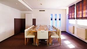 Un salón de conferencias con una larga mesa y sillas, paredes blancas y ventanas amplias con persianas.