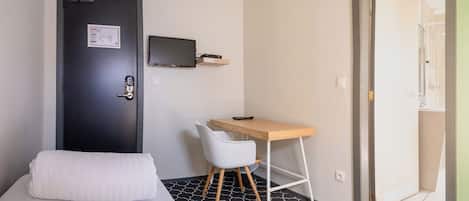 A hotel room with a bed, a desk, a chair, a TV mounted on the wall, and a door with a sign.