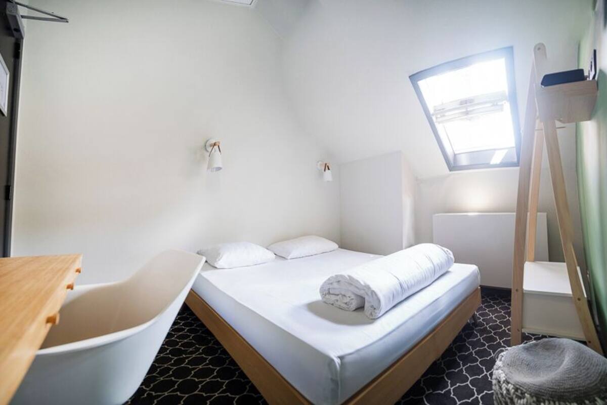 A compact hotel room with a single bed, a desk, a chair, a small table, a window with a view, and a skylight.